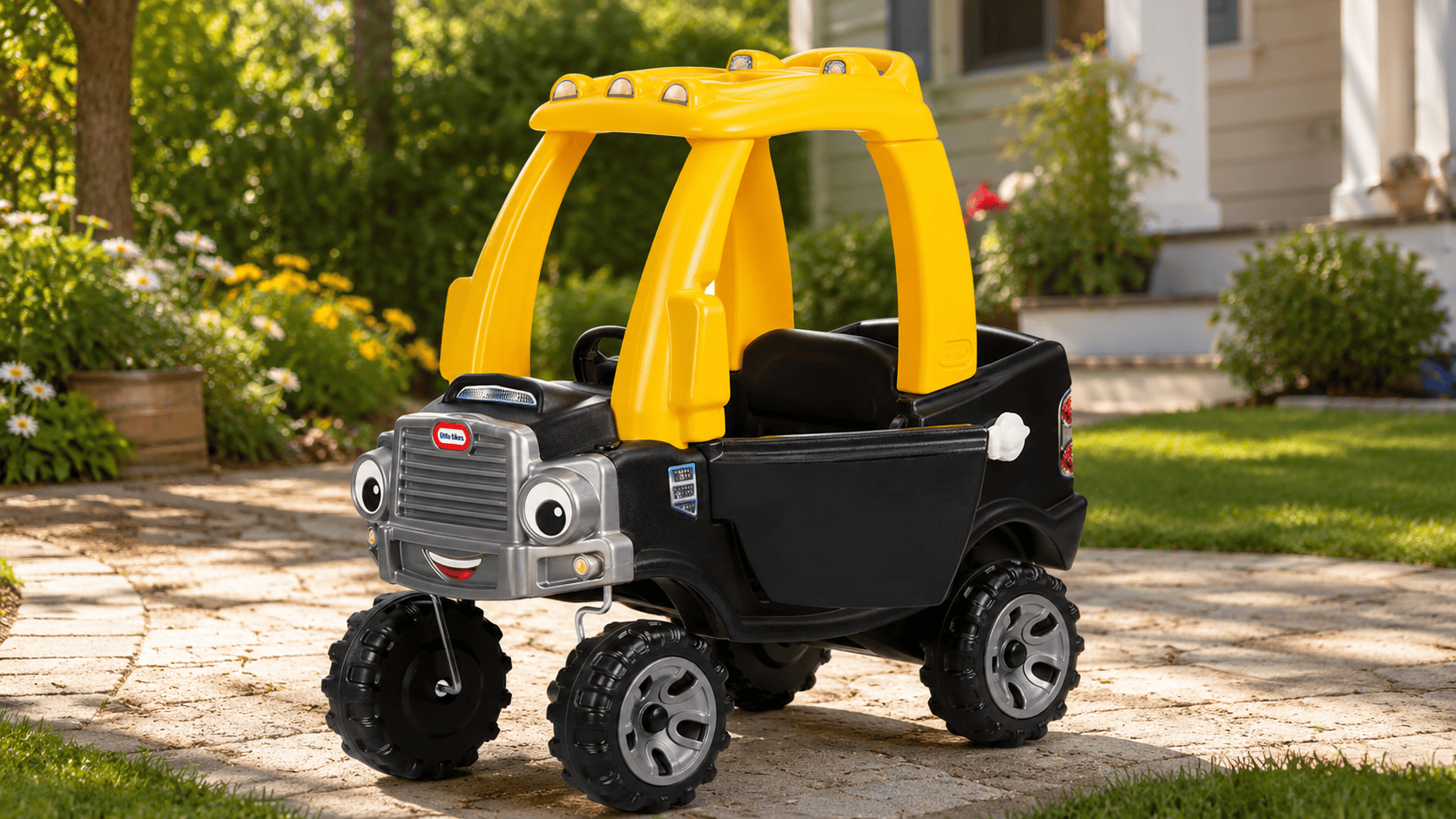 Little Tikes Cozy Truck Ride-On in black and yellow on a sunny summer driveway beside a landscaped backyard.