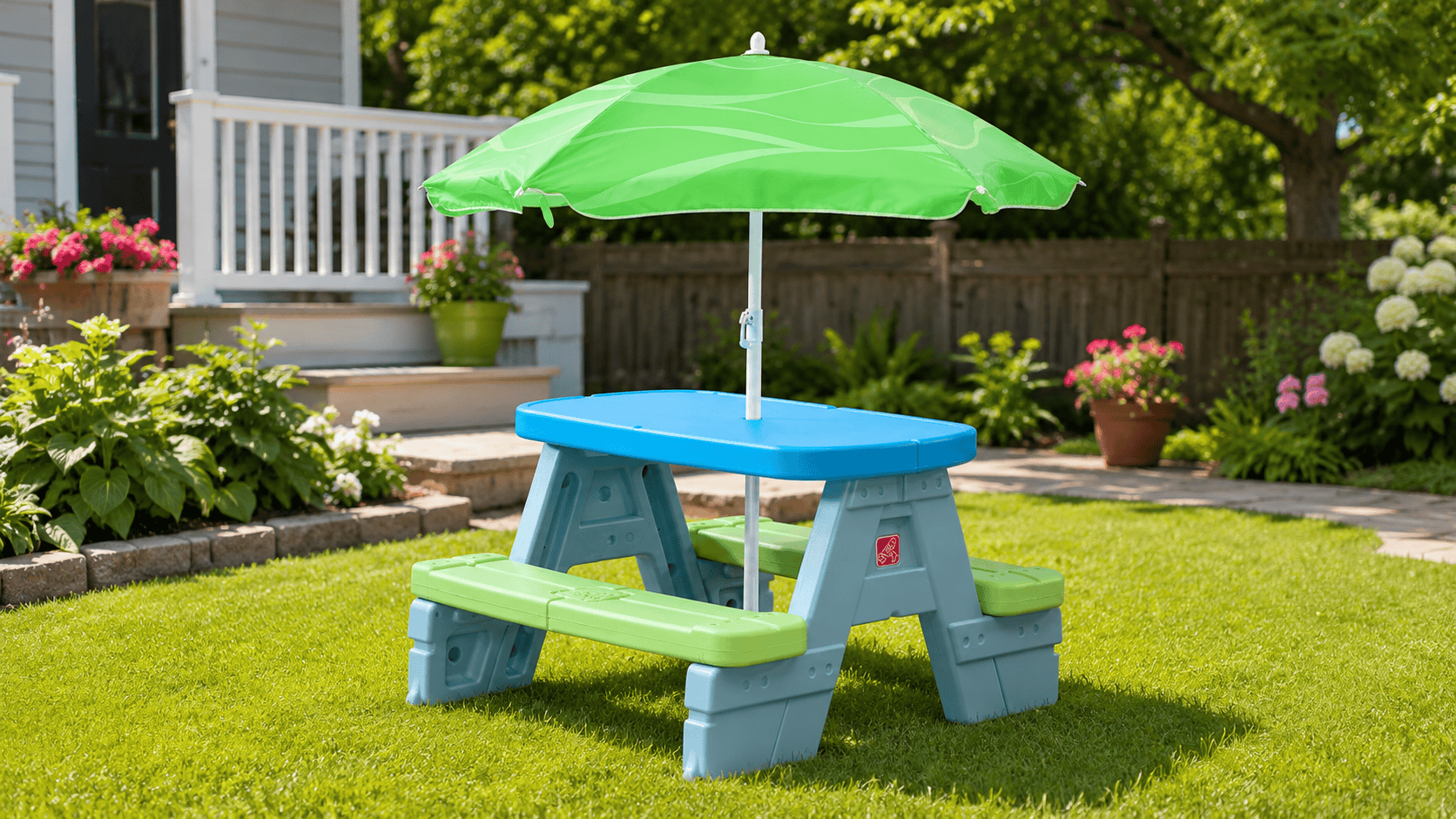 Step2 Sun & Shade Kids Picnic Table with removable umbrella set up in a sunny backyard with green grass, flowers, and a shaded outdoor play area